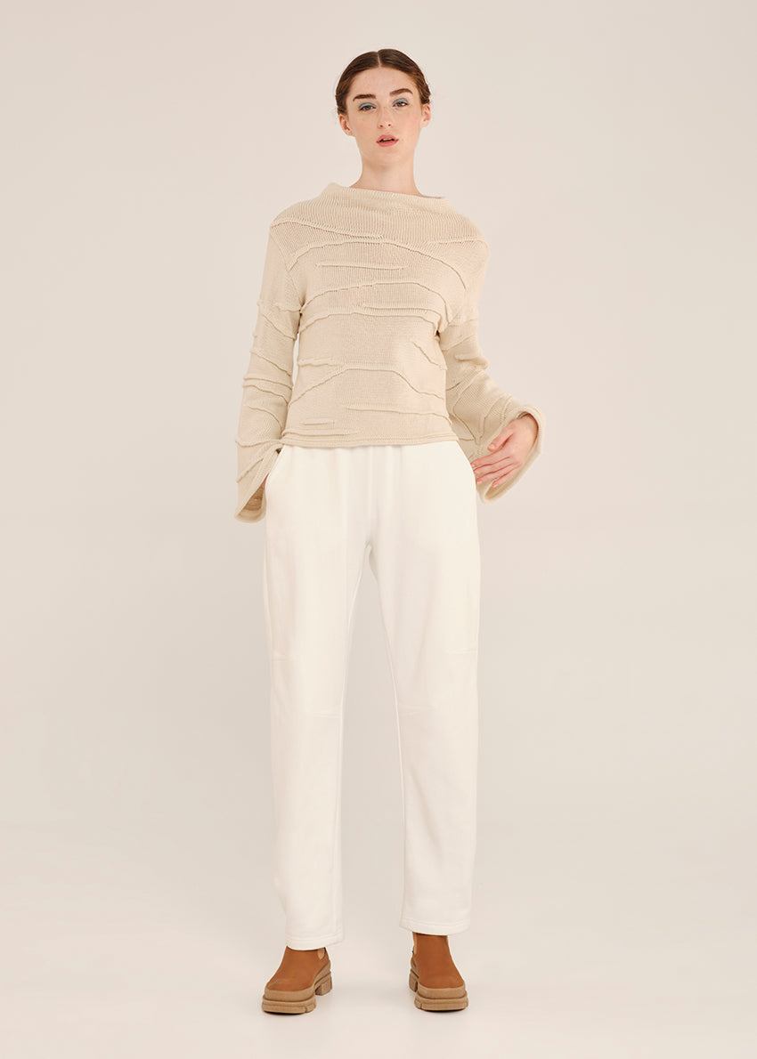 Person wearing a beige textured sweater and white pants on a neutral background