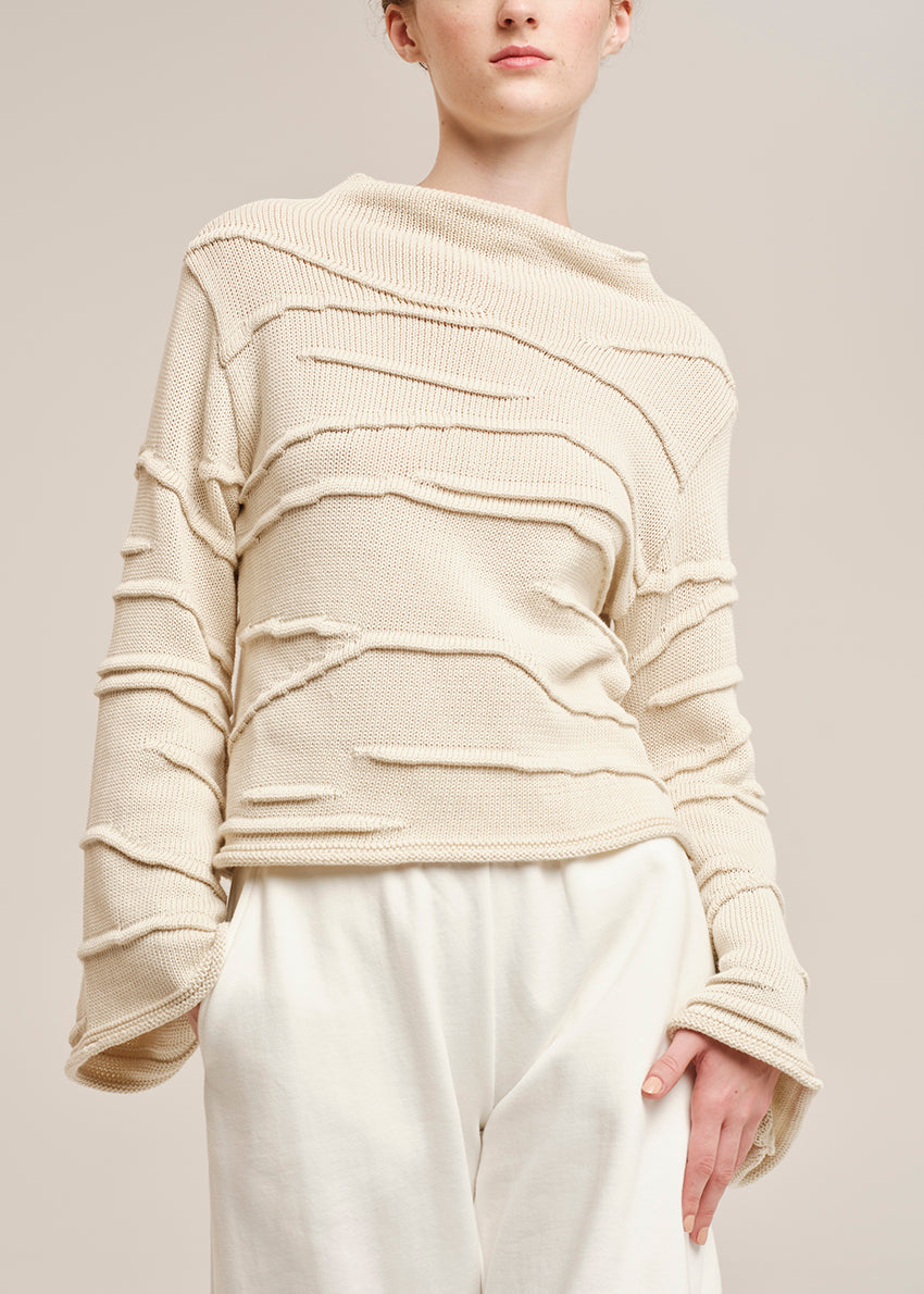 Person wearing a beige textured sweater and white pants on a neutral background