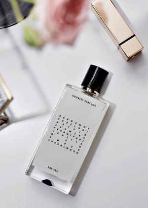 A clear glass bottle of Agonist Parfums' Say Yes Unisex Eau de Parfum with a black cap. The label lists the fragrance notes: bergamot, hazelnut, dry fig, jasmine, frangipani, vanilla, oud, amber, sandalwood.