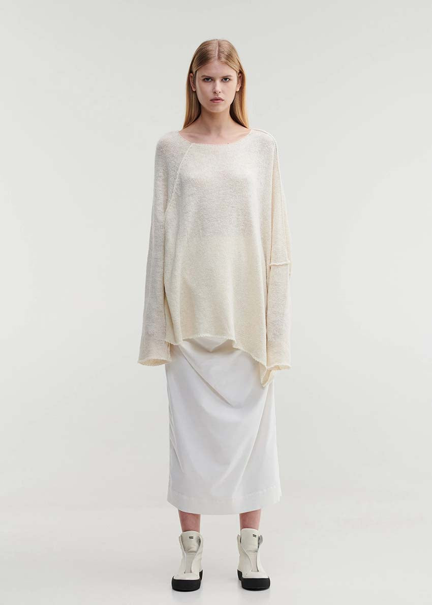 A female model wearing a beige, loosely knitted sweater with a dropped shoulder and asymmetric hemline, paired with a white skirt.