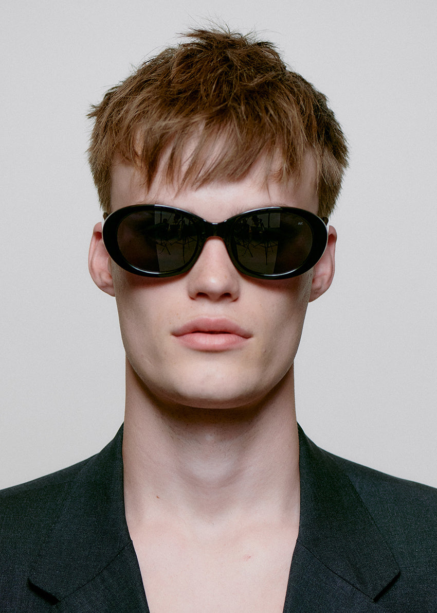 Person wearing dark sunglasses and a black jacket against a plain background