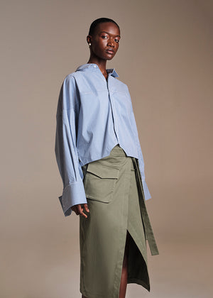 A model wearing a green wrap-style skirt with a large pocket and a front slit, paired with a blue shirt.
