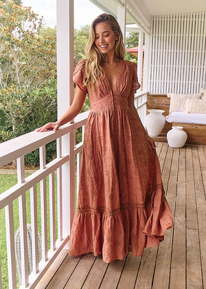 A woman standing on a porch wearing a brown, solid pattern, Maxi dress with short flutter sleeves, V-neckline, and ruffled hem.