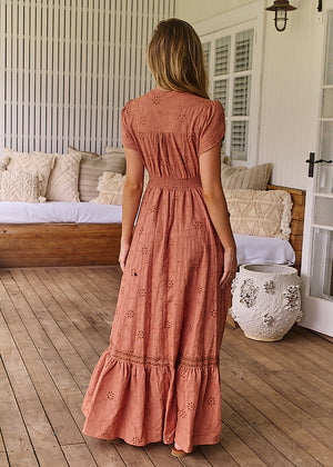 A woman standing on a porch wearing a brown, solid pattern, Maxi dress with short flutter sleeves, V-neckline, and ruffled hem.