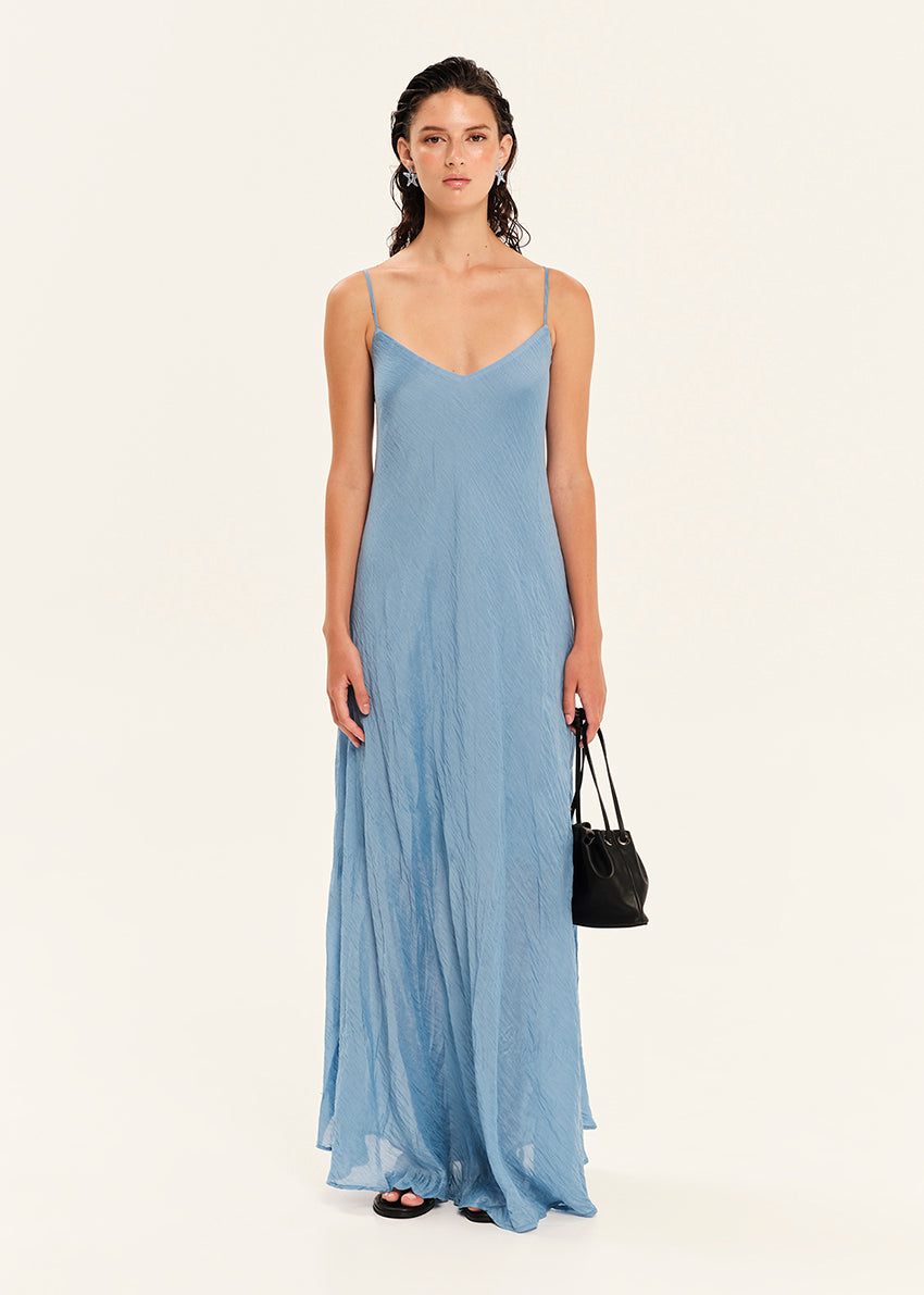 Woman wearing a long light blue dress holding a black handbag on a white background
