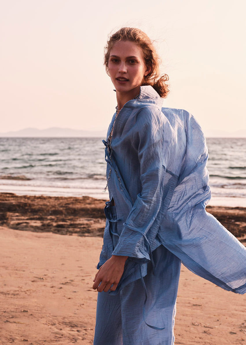 Woman in a light blue shirt with matching pants standing on a beach with ocean in the background
