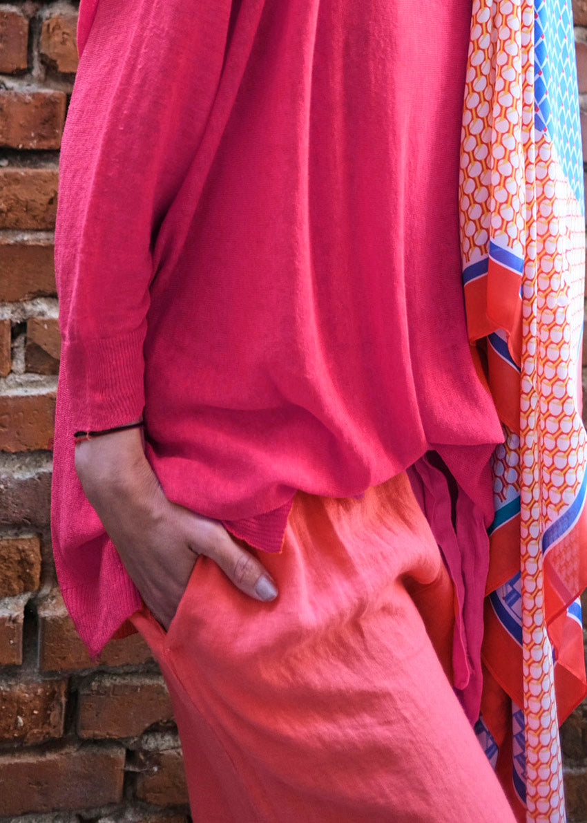 A person wearing a deep pink, V-neck, long-sleeved pullover with slits on the sides, over a patterned scarf, against a brick wall background.