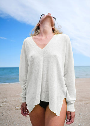 A person standing on a beach wearing a white, long-sleeved pullover with a deep V-neck, over the hips silhouette.