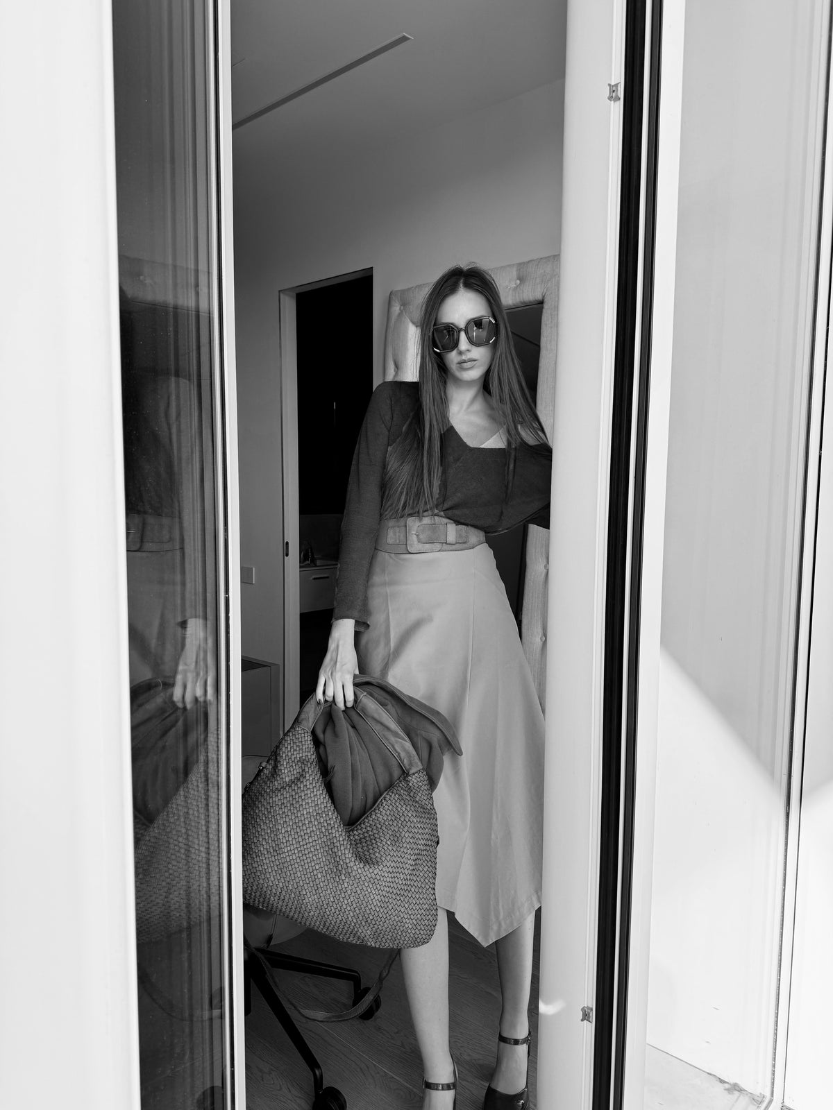 Woman in sunglasses holding a bag, standing in a doorway.