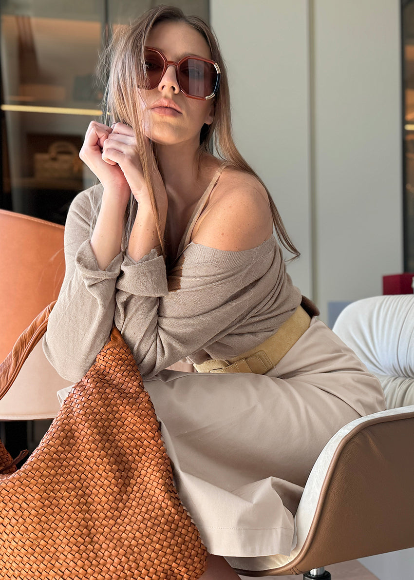 Woman sitting on a chair holding a woven bag, wearing sunglasses and a beige outfit.