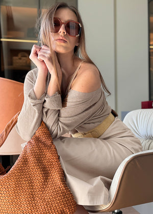 Woman sitting on a chair holding a woven bag, wearing sunglasses and a beige outfit.