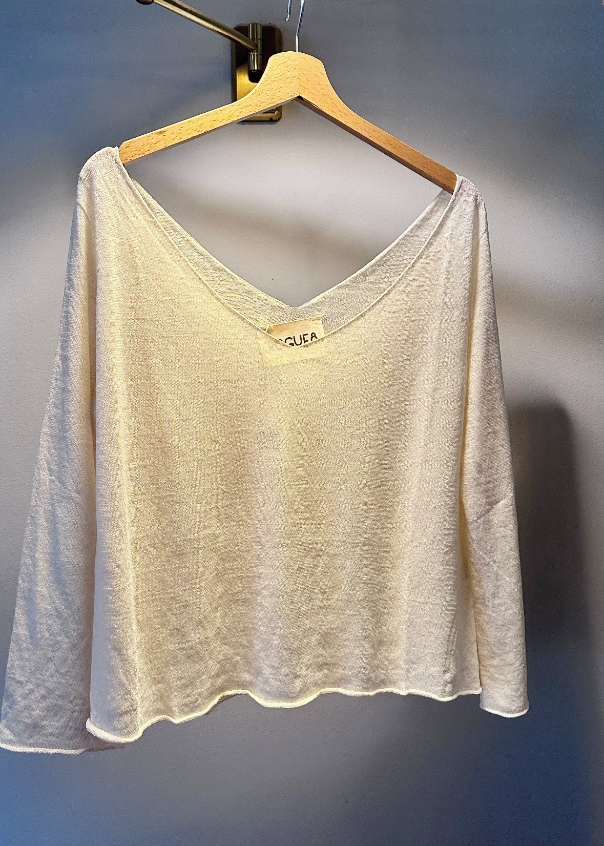A beige, linen short sleeve pullover sweater with a deep V-neckline, displayed on a wooden hanger against a blue background.