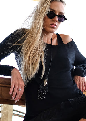 A woman sitting with long blonde hair and wearing a black sweater with long sleeves, paired with sunglasses and a necklace.