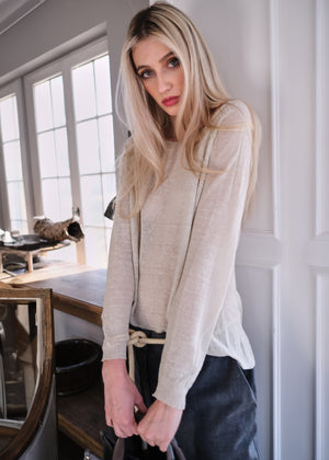 A woman wearing a beige, linen-textured sweater with long sleeves and a boat neckline, paired with black pants.