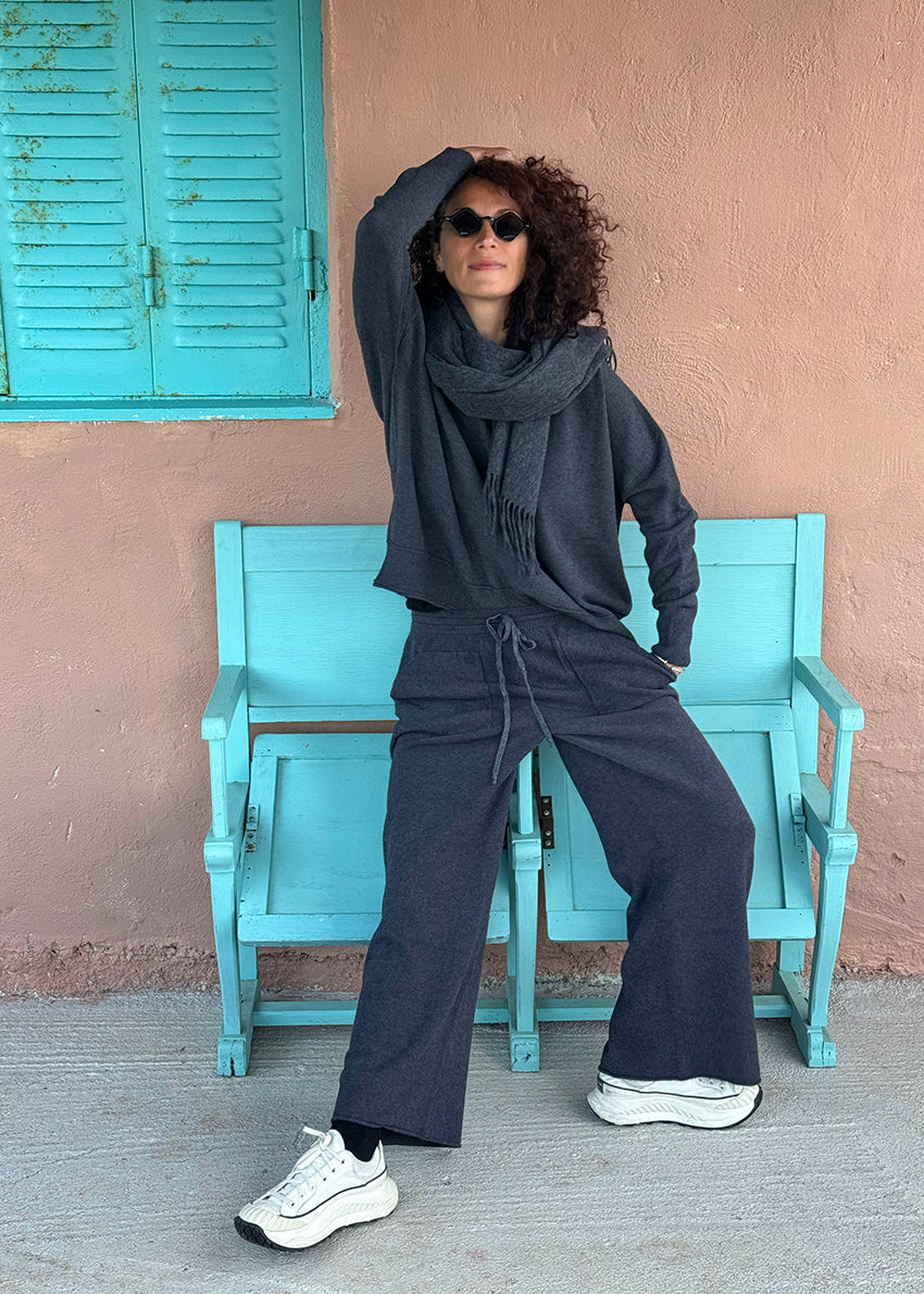 Person wearing a dark outfit with sunglasses, sitting on a blue bench against a pink wall.