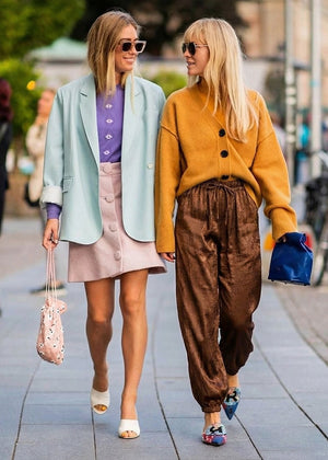Two women walking on a sidewalk, one in a light blue blazer and pink skirt, the other in a mustard yellow cardigan and brown pants.