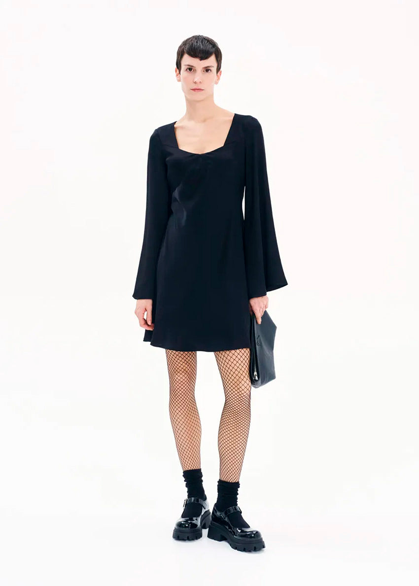 A model wearing a black mini dress with long bell sleeves and a V-neckline, standing against a white background.