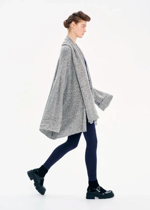 A model wearing a gray knit cardigan with long sleeves and a non-detachable scarf, paired with dark pants and black boots.