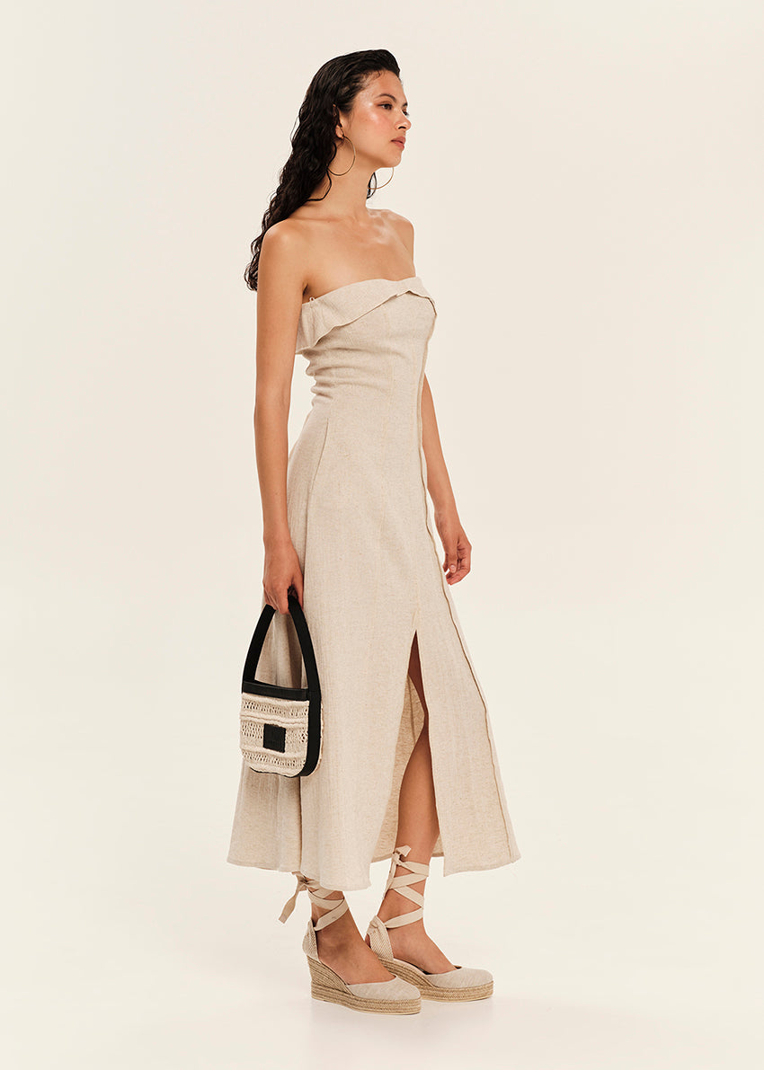Woman wearing a strapless beige dress with a slit, holding a black and white handbag on a plain background.
