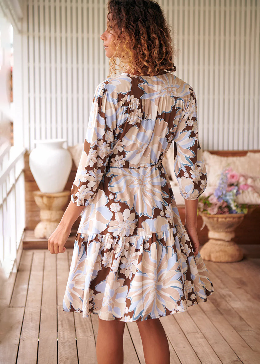 A woman standing on a porch wearing a floral mini dress with a cinch waist and three-quarter sleeves.