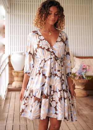 A woman standing on a porch wearing a floral mini dress with a cinch waist and three-quarter sleeves.