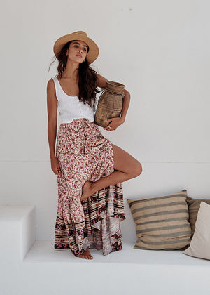A woman sitting on a bench wearing a long floral skirt, a white top, and a straw hat.