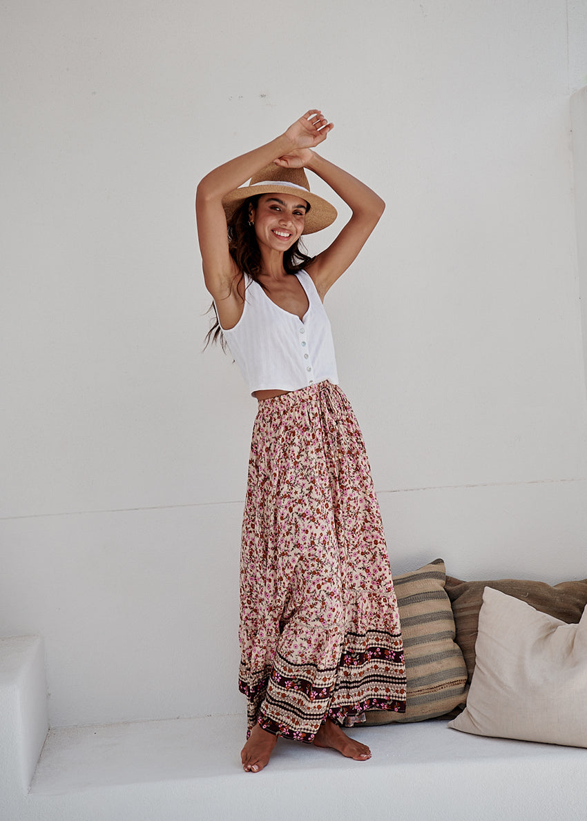 A woman sitting on a bench wearing a long floral skirt, a white top, and a straw hat.