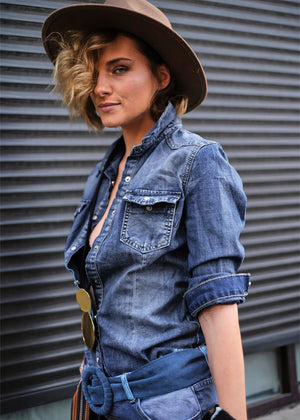 A person wearing a blue denim shirt with pockets, a brown hat, and a brown belt with a buckle, standing in front of a dark background.