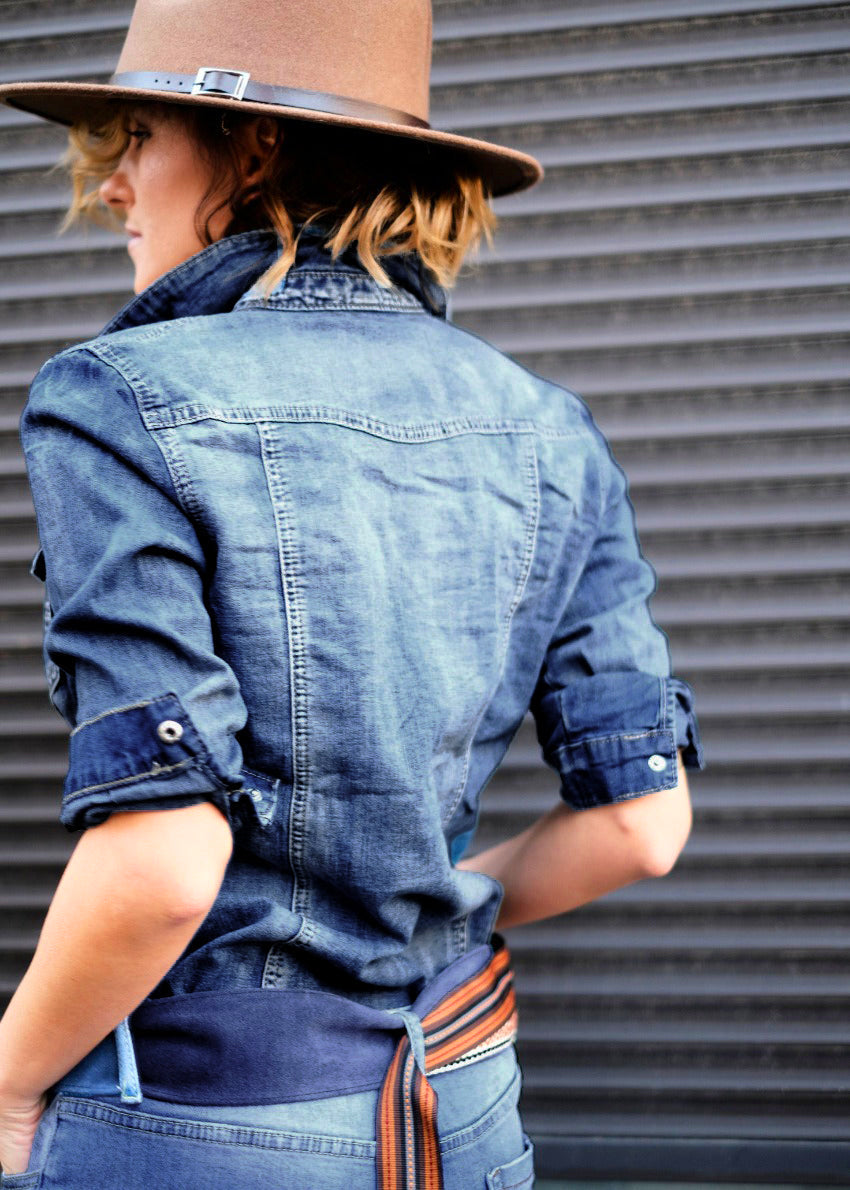 A person wearing a blue denim shirt with pockets, a brown hat, and a brown belt with a buckle, standing in front of a dark background.