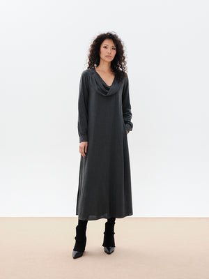 A woman standing, wearing a long gray dress with a draped cowl neck and fabric buttons on the sleeves. The dress has a comfortable fit and side openings at the hemline.