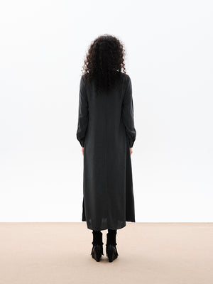 A woman standing, wearing a long gray dress with a draped cowl neck and fabric buttons on the sleeves. The dress has a comfortable fit and side openings at the hemline.