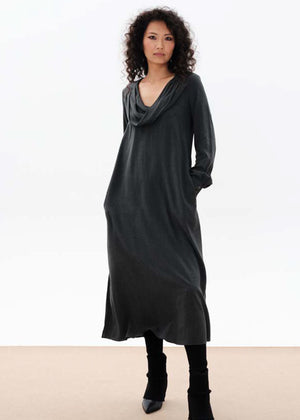 A woman standing, wearing a long gray dress with a draped cowl neck and fabric buttons on the sleeves. The dress has a comfortable fit and side openings at the hemline.