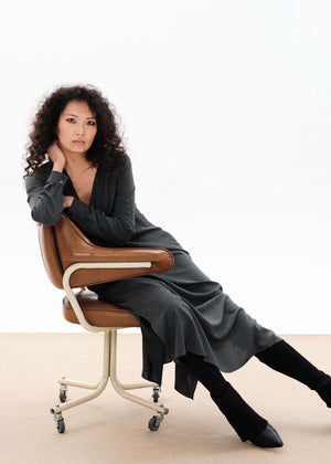 A woman sitting, wearing a long gray dress with a draped cowl neck and fabric buttons on the sleeves. The dress has a comfortable fit and side openings at the hemline