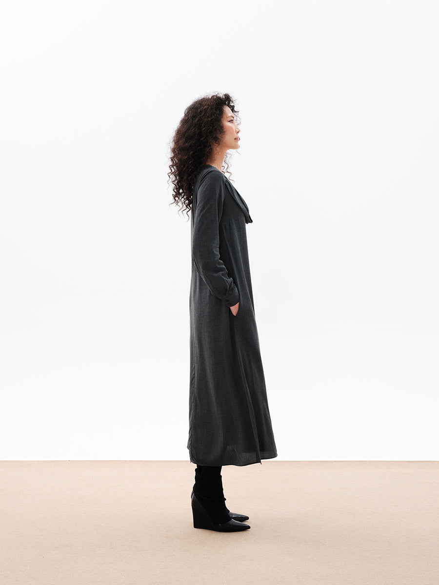 A woman standing, wearing a long gray dress with a draped cowl neck and fabric buttons on the sleeves. The dress has a comfortable fit and side openings at the hemline.