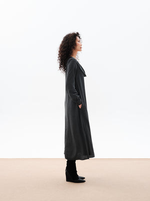 A woman standing, wearing a long gray dress with a draped cowl neck and fabric buttons on the sleeves. The dress has a comfortable fit and side openings at the hemline.