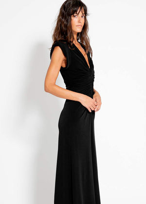 A woman wearing a long black dress with a V-neckline and knotted waistline, standing against a plain background.