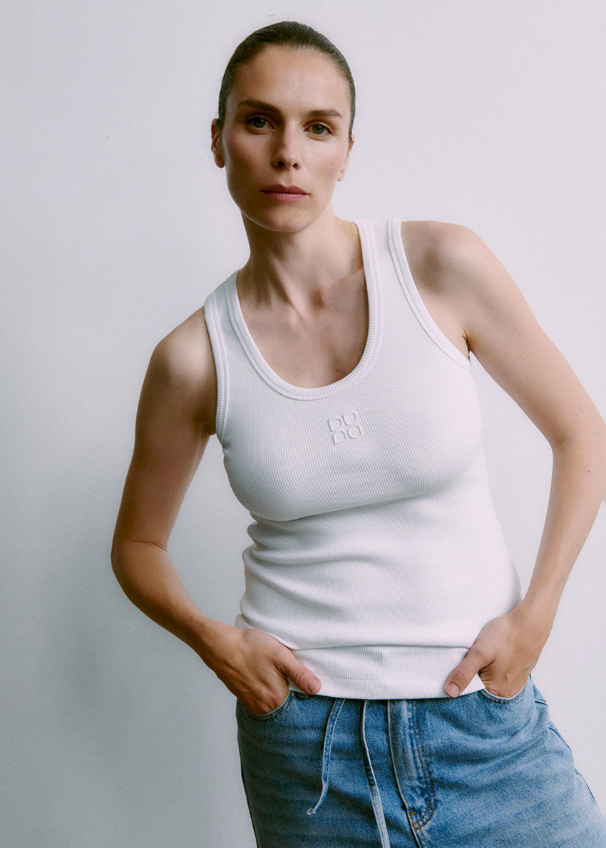 Person wearing a white tank top with a logo and blue jeans against a plain background