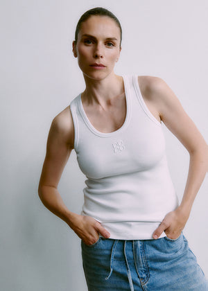 Person wearing a white tank top with a logo and blue jeans against a plain background