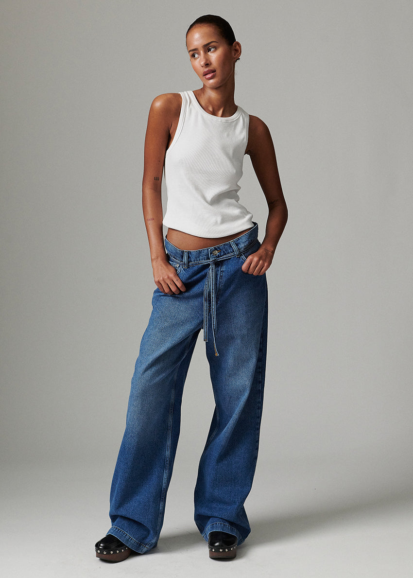 Woman wearing a white tank top and blue jeans against a gray background