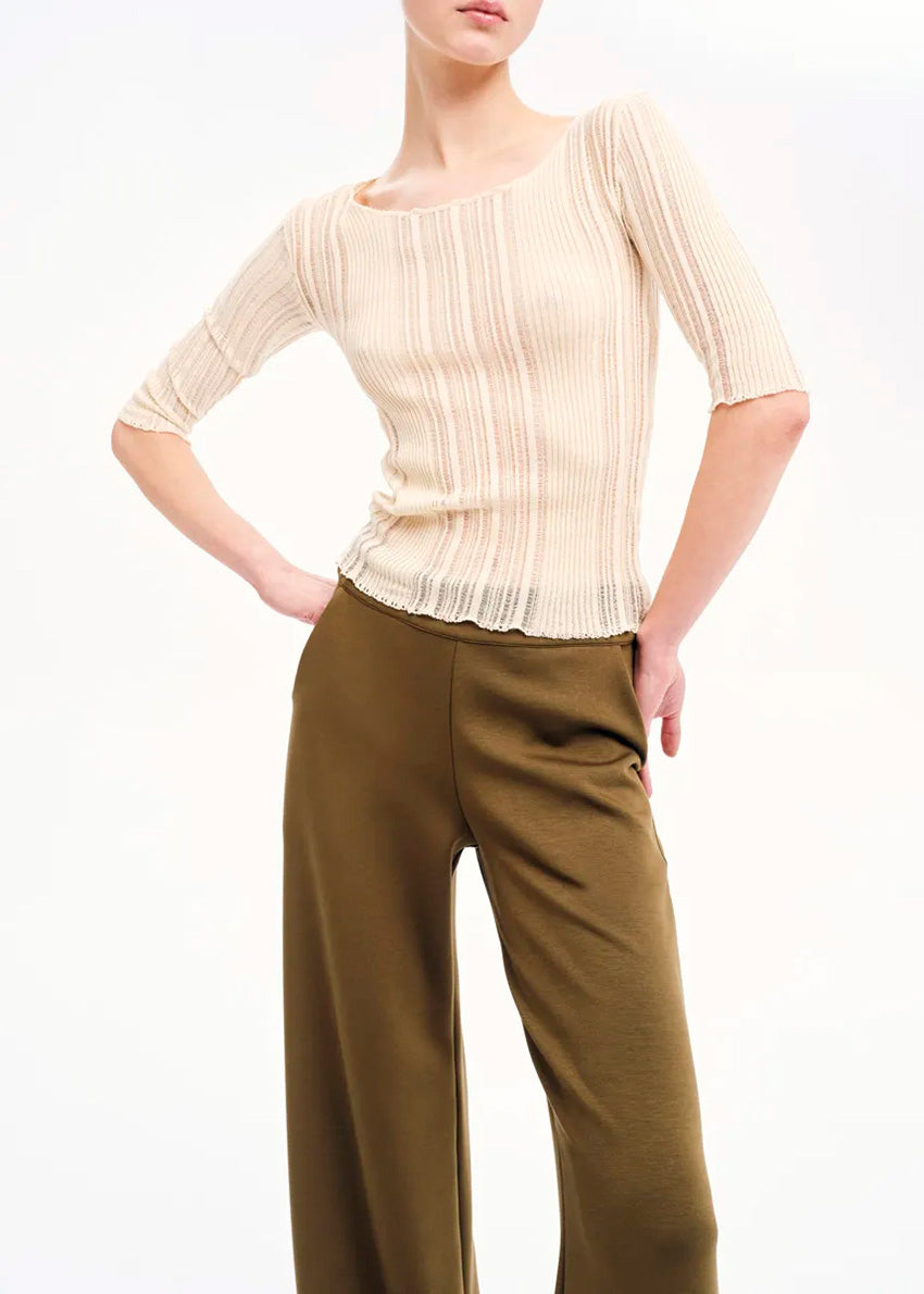 A woman modeling a beige knitted top with rib pattern and 3/4 sleeves, paired with green trousers.