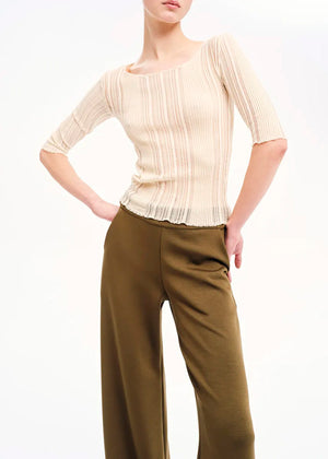 A woman modeling a beige knitted top with rib pattern and 3/4 sleeves, paired with green trousers.