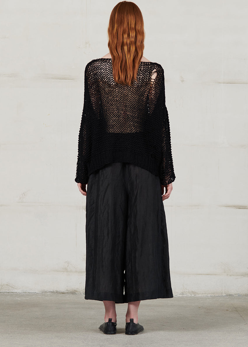 Person wearing a black textured top and wide-leg pants against a white wall.