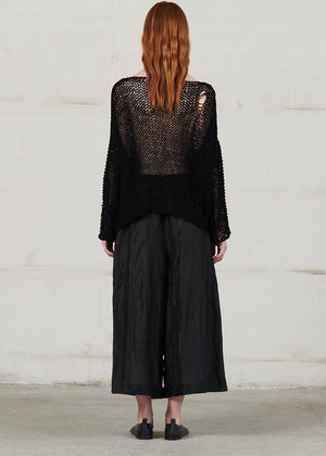 Person wearing a black textured top and wide-leg pants against a white wall.