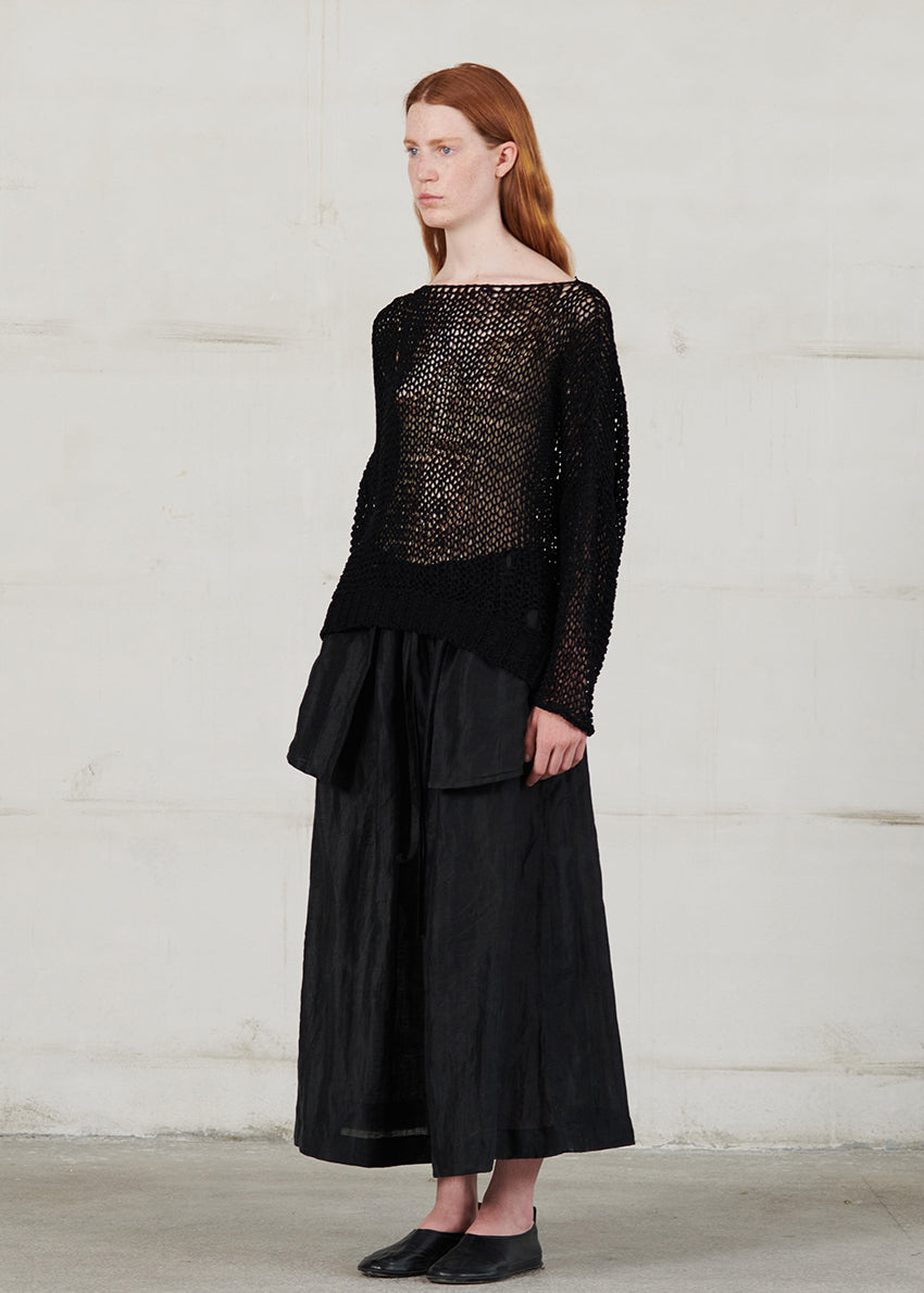 Woman wearing a black outfit with a textured top and flowing skirt against a white wall.