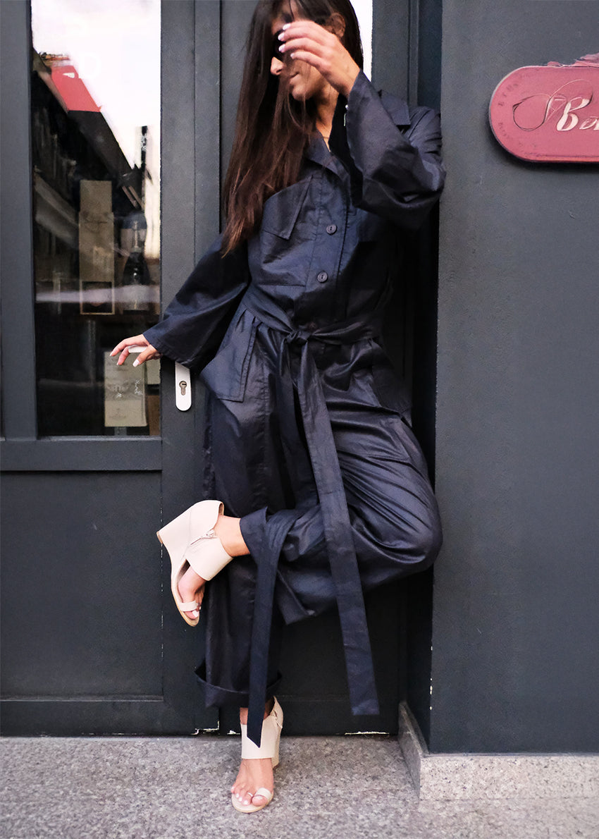 Woman wearing a jumpsuit standing against a dark wall.
