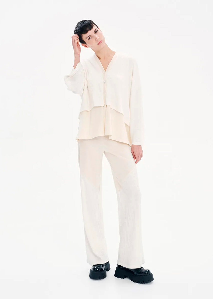 A person standing on a sidewalk wearing high-waisted white trousers and a white top, with their hands in their pockets.