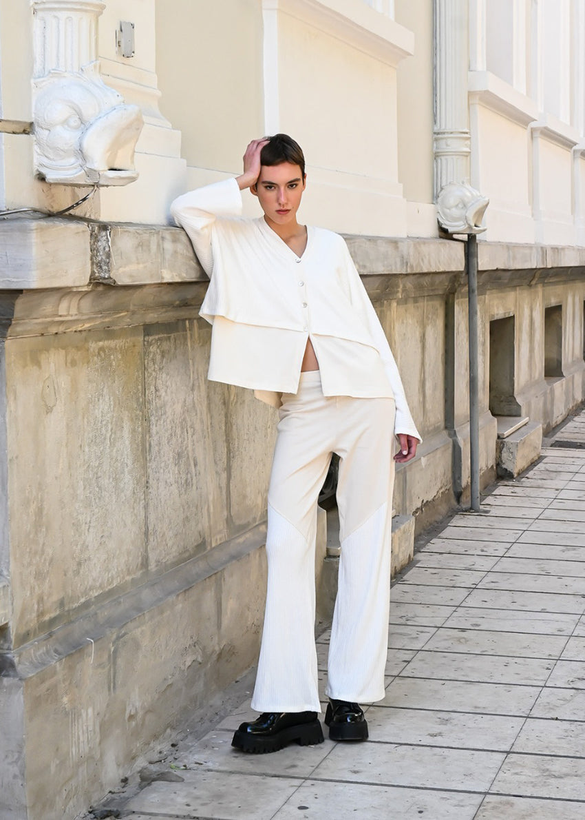 A person standing on a sidewalk wearing high-waisted white trousers and a white top, with their hands in their pockets.