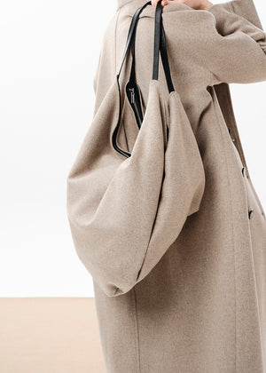 A person wearing a beige coat is holding a beige oversized slouchy double bag with a zipper closure and a shoulder strap.