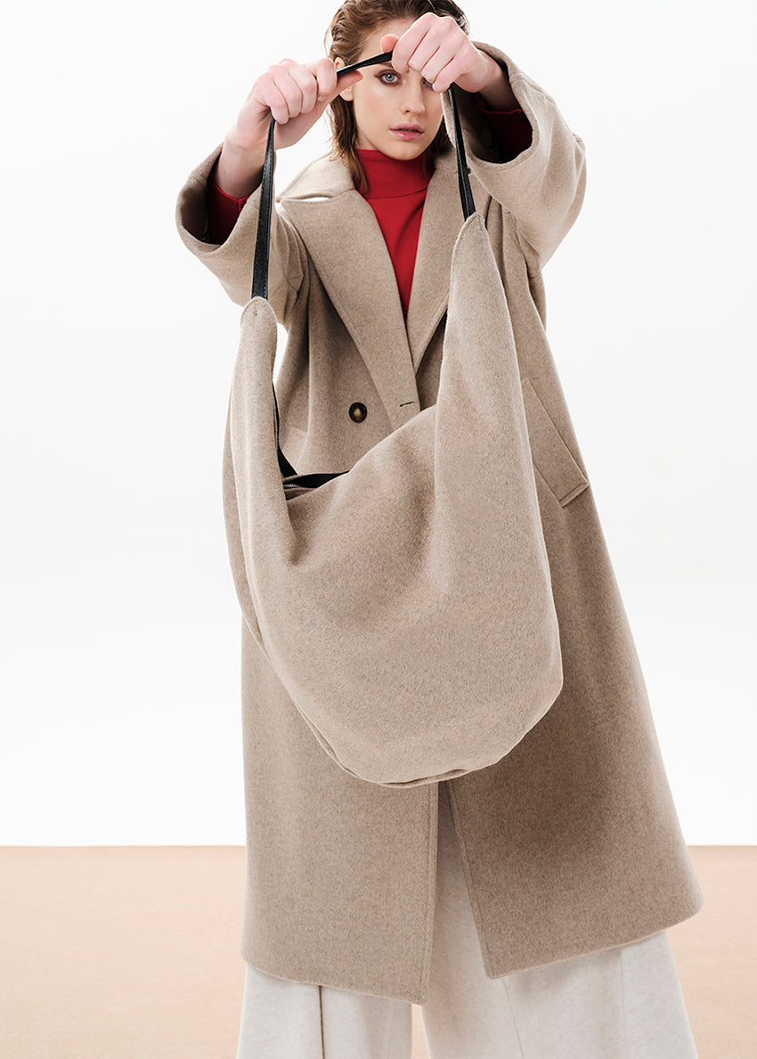 A person wearing a beige coat is holding a beige oversized slouchy double bag with a zipper closure and a shoulder strap.