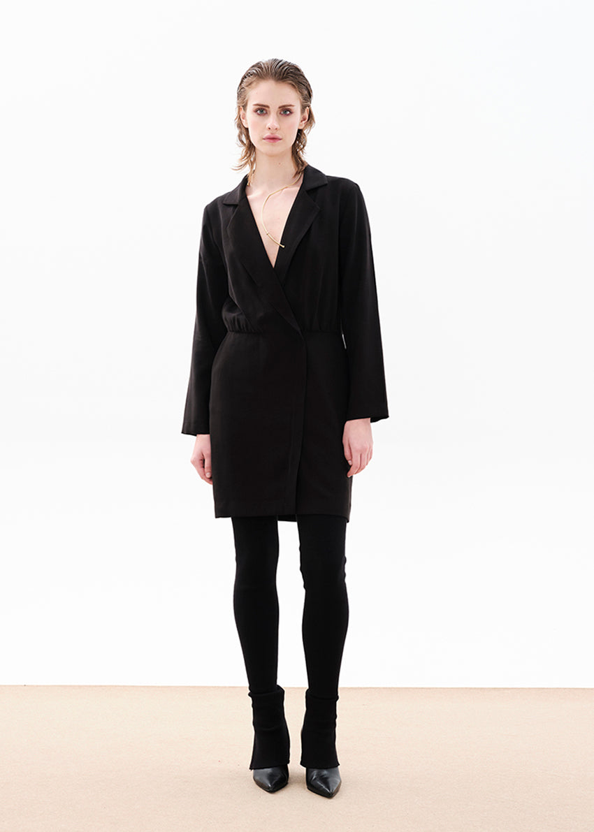 A female model wearing a black draped blazer dress with a V-neck collar, long sleeves, and a two-way zipper front.